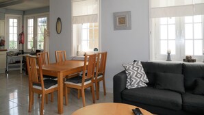 Un'area pranzo con tavolo e sedie in legno, un divano nero con cuscini decorati e ampie finestre con tende bianche.