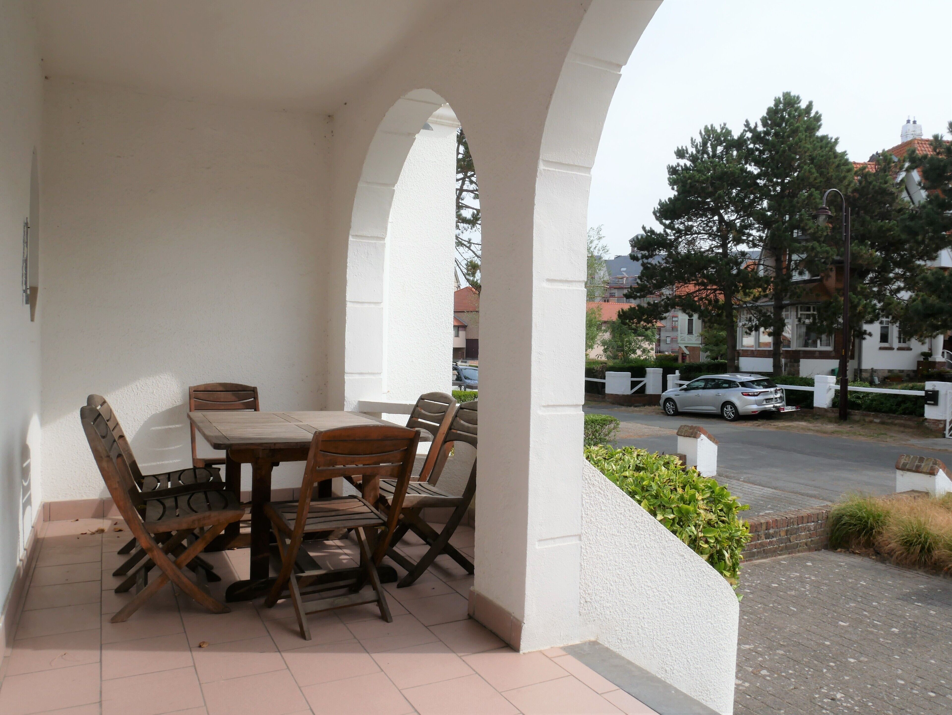 Un espace extérieur abrité, avec une table et des chaises, un mur blanc et une vue sur la rue et les bâtiments en arrière-plan.
