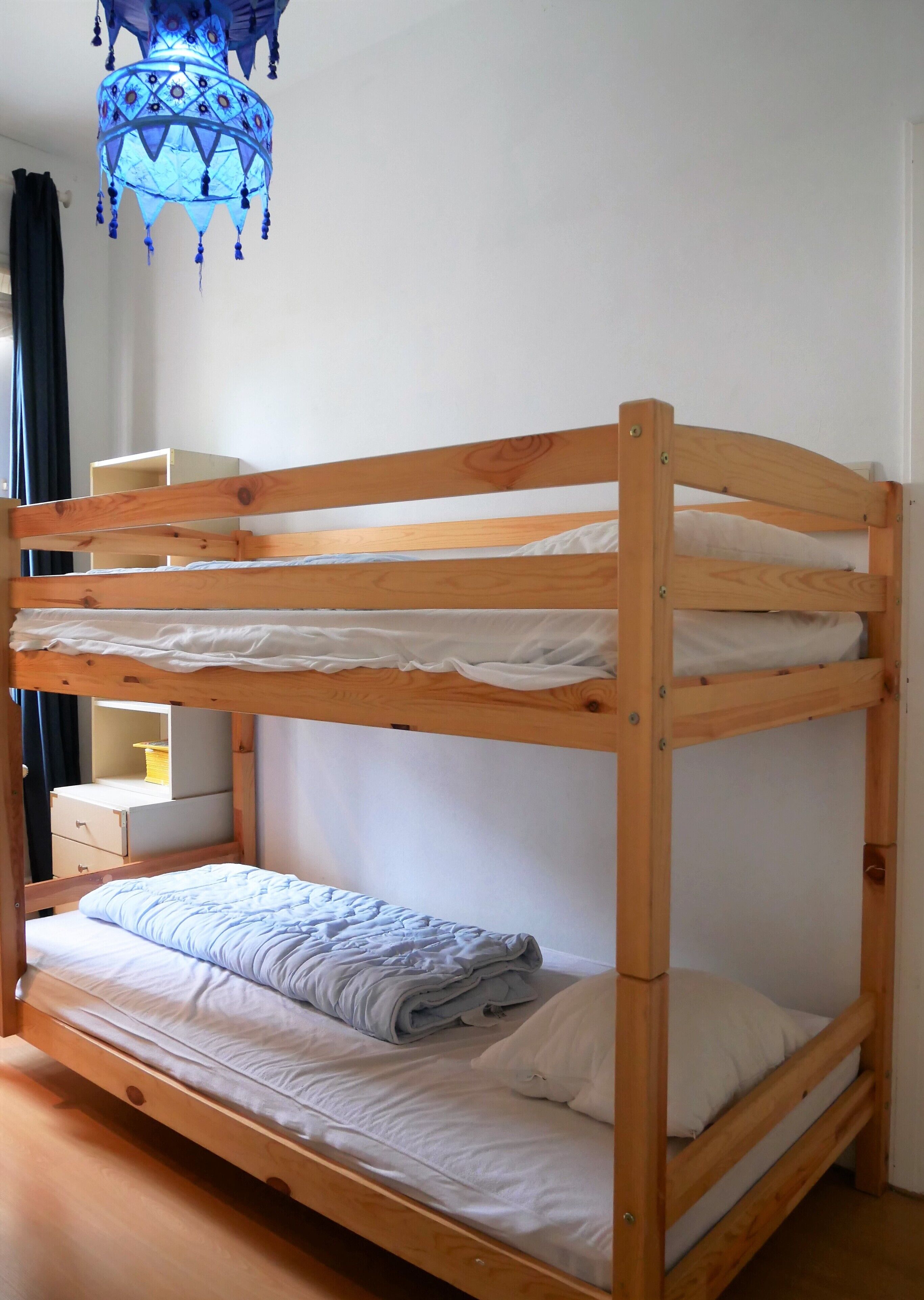 Una cama litera con estructura de madera, ropa de cama blanca y una lámpara colgante azul arriba.