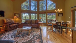 A spacious living room with a large window, a fireplace, a dining area, and a cozy seating area. - Sable Pine Cabin | Sleeps 8 | Nature Trails | Wood Stove (Area F (Columbia Lake/Windermere Lake))