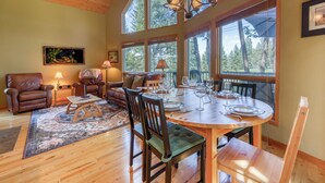 A cozy living room with a wooden ceiling, large windows, a dining table set for four, and a leather sofa. - Sable Pine Cabin | Sleeps 8 | Nature Trails | Wood Stove (Area F (Columbia Lake/Windermere Lake))