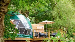 A wooden cabin with a glass dome structure, a deck with chairs and an umbrella, surrounded by dense greenery. - Chalet in Kent with Hot Tub & Countryside Views (Hoath)
