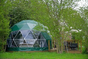 Relax in a geodesic dome tent surrounded by lush greenery, with a transparent window and adjacent deck for a peaceful retreat.