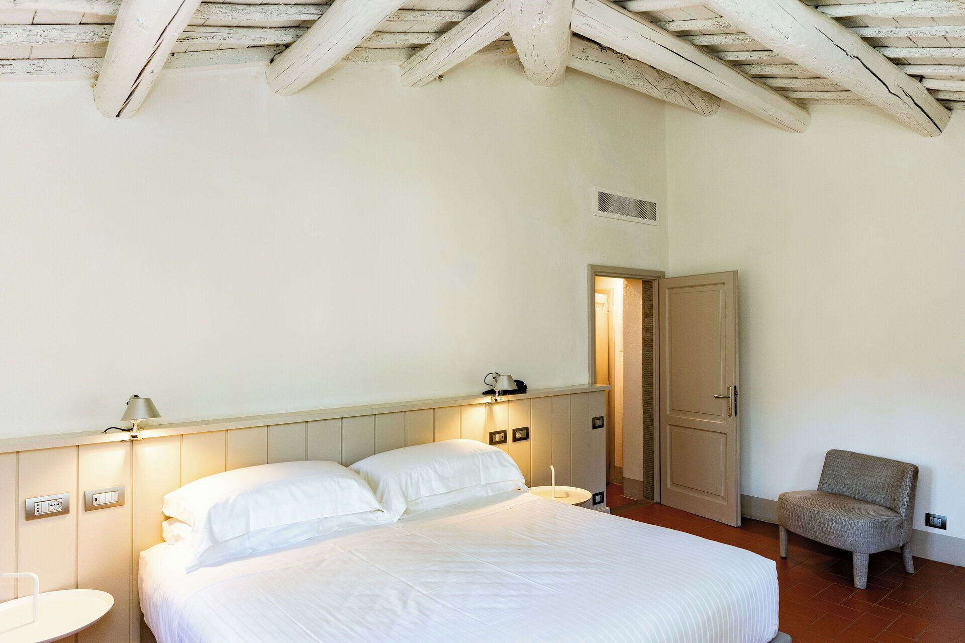 Een slaapkamer met een bed, bedlampen, een stoel en een deur naar een andere kamer.