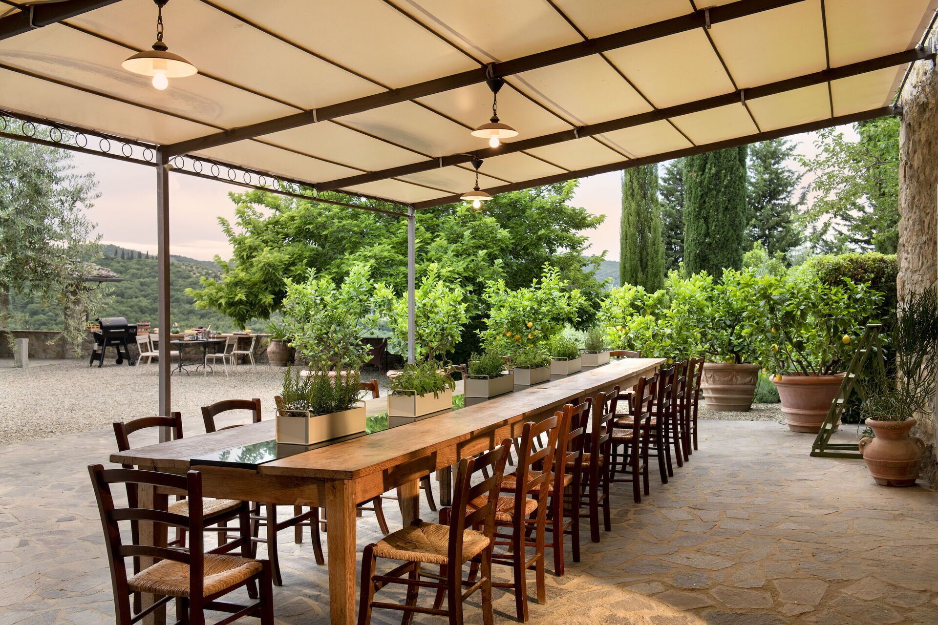 An outdoor patio with a long wooden table and chairs, surrounded by potted plants and a view of a lush landscape.