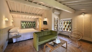 Une pièce comprenant un lit, un canapé vert, une table basse en bois avec un livre, un fauteuil en osier et une fenêtre ornée d’un motif en treillis.