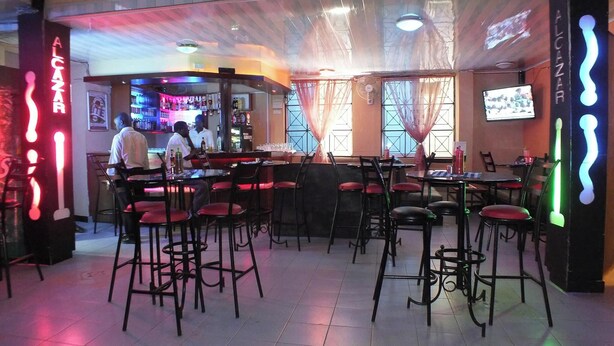A bar area with stools and tables, a counter with a bartender, and a television screen on the wall. - Alcazar Hotel Kisumu (Kisumu)