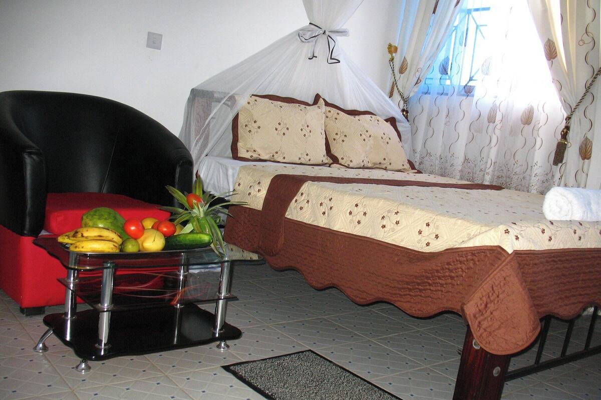 Une chambre à coucher avec un lit, une chaise, une table avec des fruits et une fenêtre avec des rideaux.