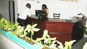 A reception desk with a computer monitor, framed certificates on the wall, and potted plants in the foreground. - Alcazar Hotel Kisumu (Kisumu)