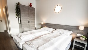 A double bed with white linens, a wooden wardrobe, a bedside table with a lamp, and a wall-mounted light fixture.