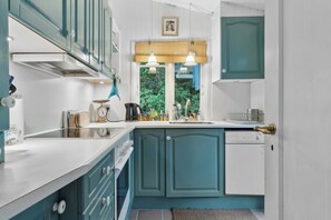 A compact kitchen with teal cabinetry, a white countertop, a window with a view of greenery, and a door with a brass handle.