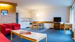 A modern hotel room with a wooden accent wall, a sofa, a red armchair, a dining table, and a kitchen area. - Appartement im Panoramic Hotel Bad Lauterberg (Bad Lauterberg)