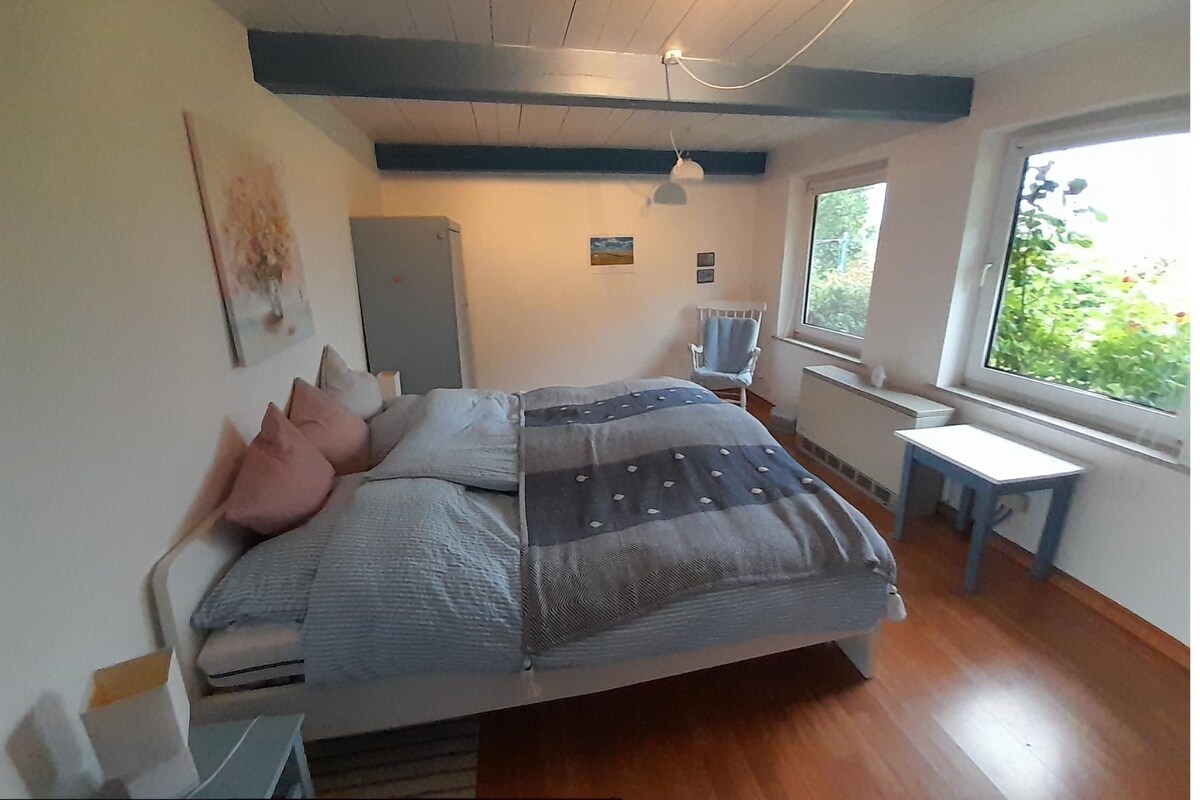 A bedroom with a bed, a desk, a chair, a radiator, and a window with a view of greenery.