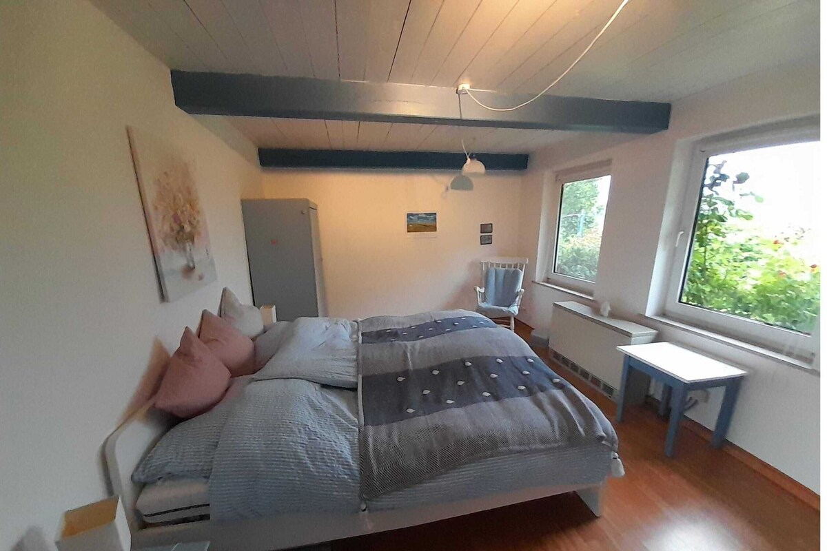 Une chambre à coucher comprenant un lit, un bureau, une chaise, un radiateur et une fenêtre donnant sur un espace verdoyant.