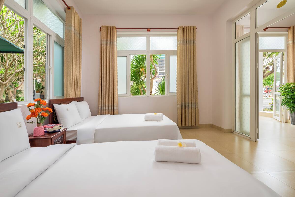 Habitación de hotel con dos camas, una ventana con cortinas, una mesita de noche con un florero, y vista a áreas verdes.