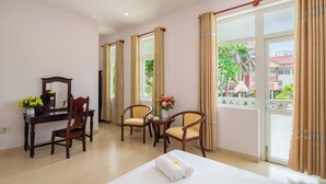 A hotel room with a bed, a desk with a mirror, two chairs, a small table with flowers, and large windows with curtains. - Villas De Mykhe (Da Nang)