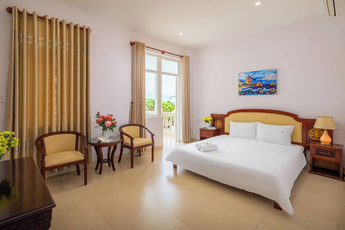 Habitación de hotel con una cama grande, dos sillas, una mesita con flores, un cuadro en la pared y una ventana con cortinas.