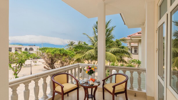 A balcony with two wooden chairs, a small table with a vase of flowers, and a view of a beach and palm trees. - Villas De Mykhe (Da Nang)