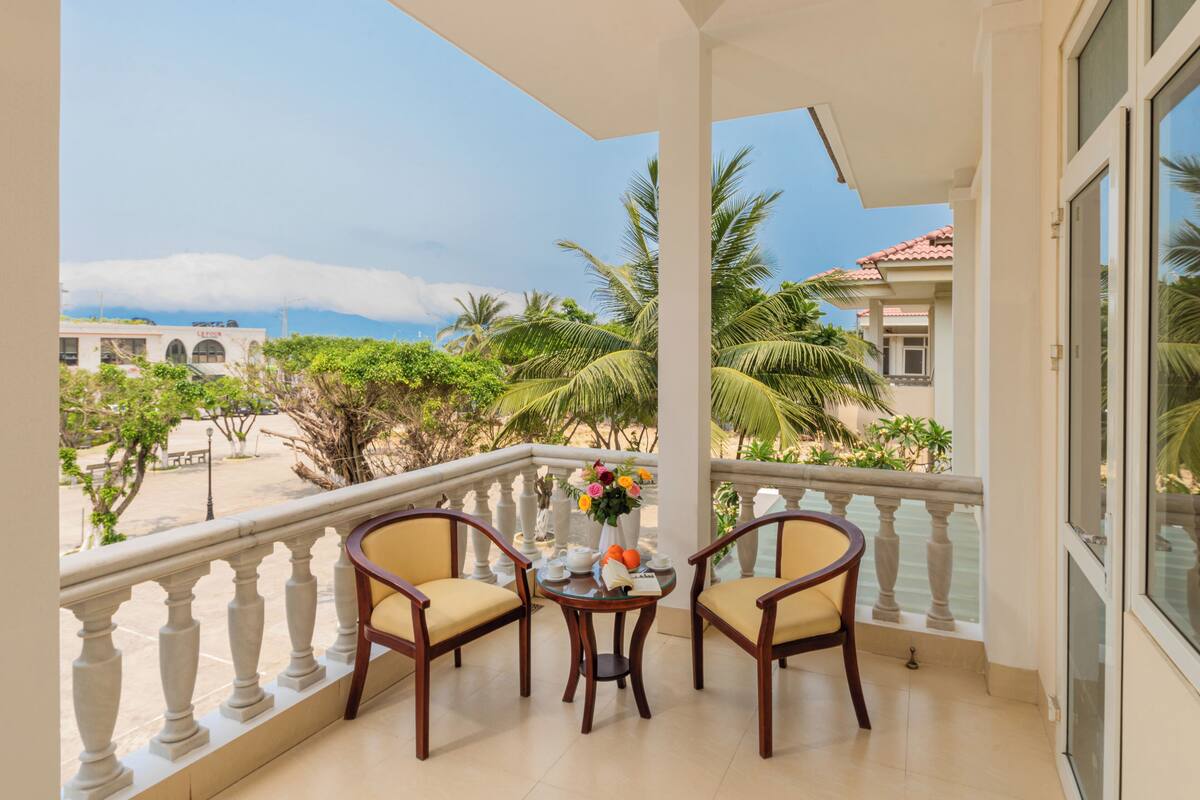 Un balcón con dos sillas de madera, una mesita con un florero y vista a una playa y palmeras.