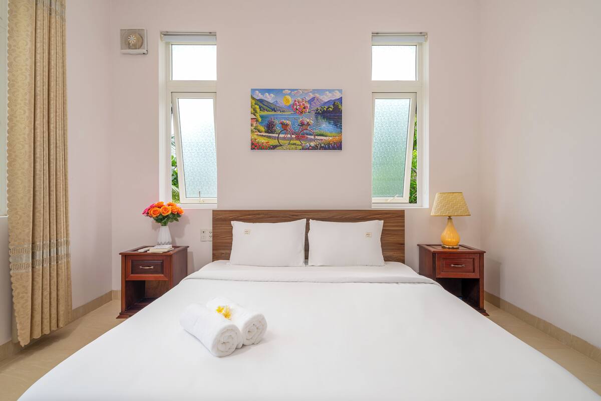 Habitación de hotel con una cama grande, dos mesitas de noche, un buró, un cuadro en la pared y una ventana con cortinas.