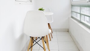Un couloir étroit aux murs blancs, avec un sol carrelé et une fenêtre sur le côté droit. On y trouve trois chaises blanches aux pieds en bois et une petite plante posée sur une table blanche.