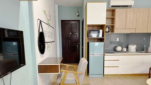 Une pièce de taille modeste comprenant un bureau en bois, une chaise, un téléviseur, un climatiseur mural, un four à micro-ondes, un réfrigérateur et un petit coin cuisine avec un évier et une bouilloire.