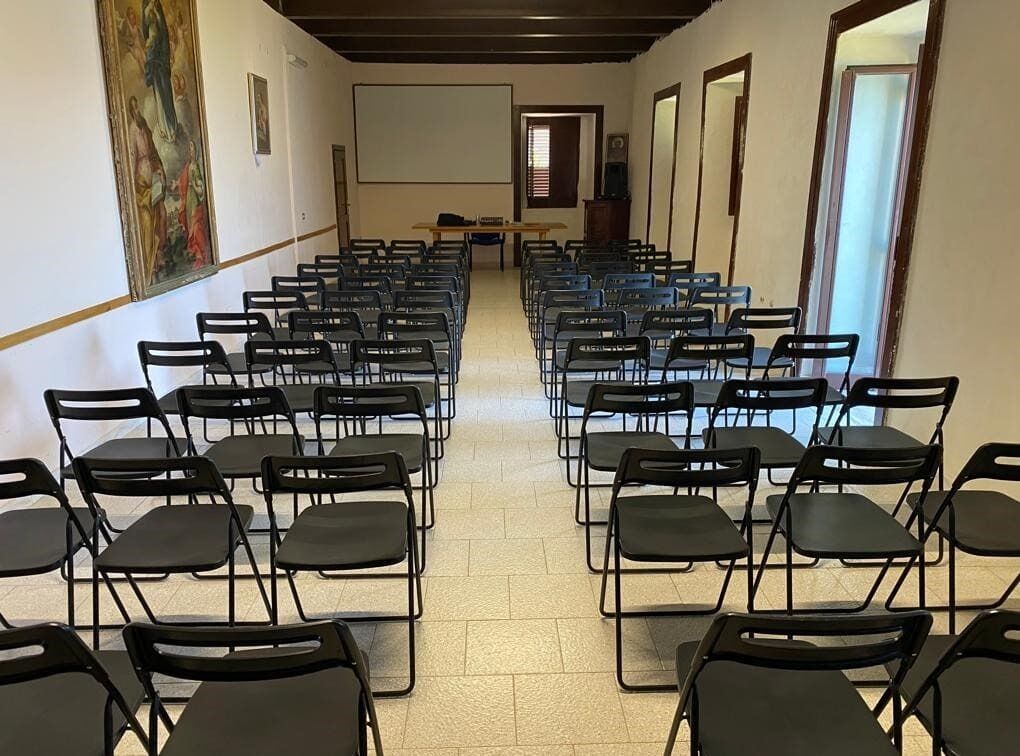 A conference room with rows of chairs facing a screen, a wooden table, and a painting on the wall.