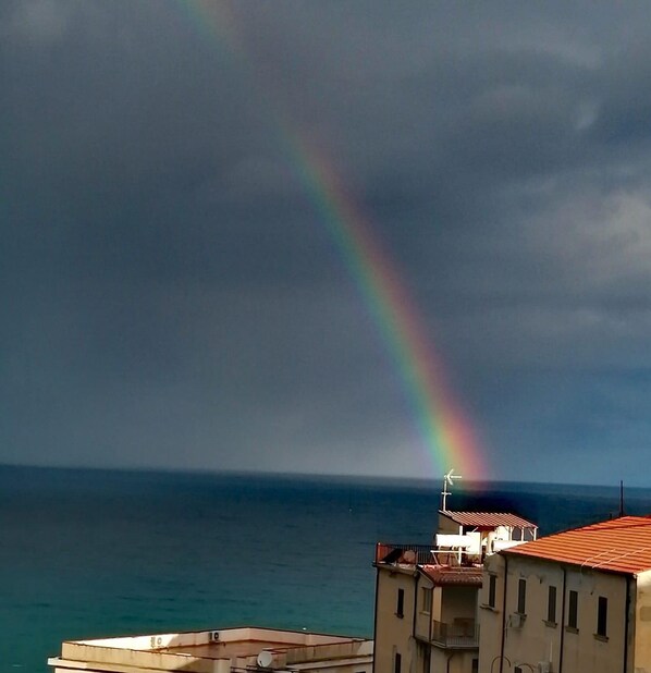 Une vue côtière avec un arc-en-ciel au-dessus de la mer, des bâtiments au premier plan et une éolienne.