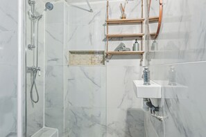 Un baño con ducha envuelta en vidrio, baldosas de mármol, estantes de madera con artículos de aseo y un lavamanos blanco.
