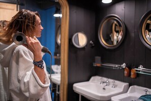 Une personne vêtue d’une robe, tenant un microphone dans une salle de bain équipée de deux lavabos, de miroirs et d’articles de toilette fixés au mur.