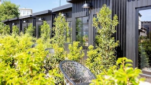 Un bâtiment moderne aux murs extérieurs sombres et aux grandes fenêtres, entouré d’une végétation luxuriante et doté d’un espace extérieur aménagé pour s’asseoir.