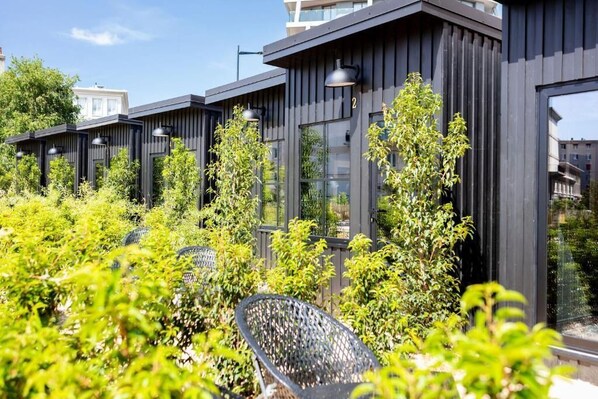 Un edificio moderno con paredes exteriores oscuras y ventanas amplias, rodeado de abundante vegetación y mobiliario para exteriores.
