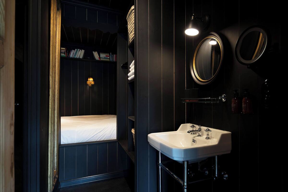 Une salle de bain à l’ambiance sombre, avec un lavabo blanc, deux miroirs et une étagère près du lit où sont posés des livres.