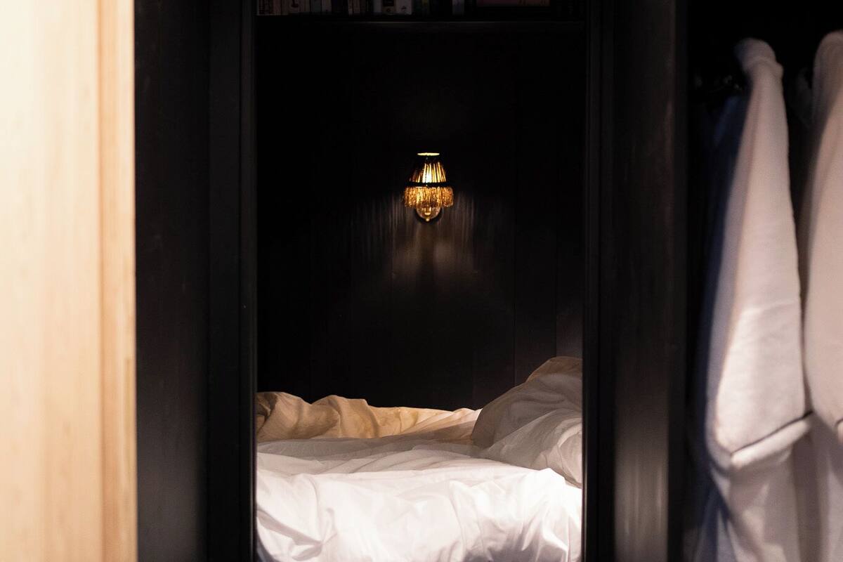 Une pièce faiblement éclairée, avec un lit, une bibliothèque et un luminaire fixé au mur.
