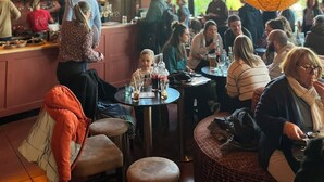 Un café animé, avec des tables et des chaises rondes, une enseigne au néon indiquant « Le Préau », et un grand luminaire suspendu.