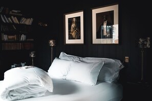 Un lit bien fait, avec du linge de lit blanc, dans une pièce à la lumière tamisée, où l’on aperçoit une bibliothèque et des tableaux encadrés accrochés au mur.