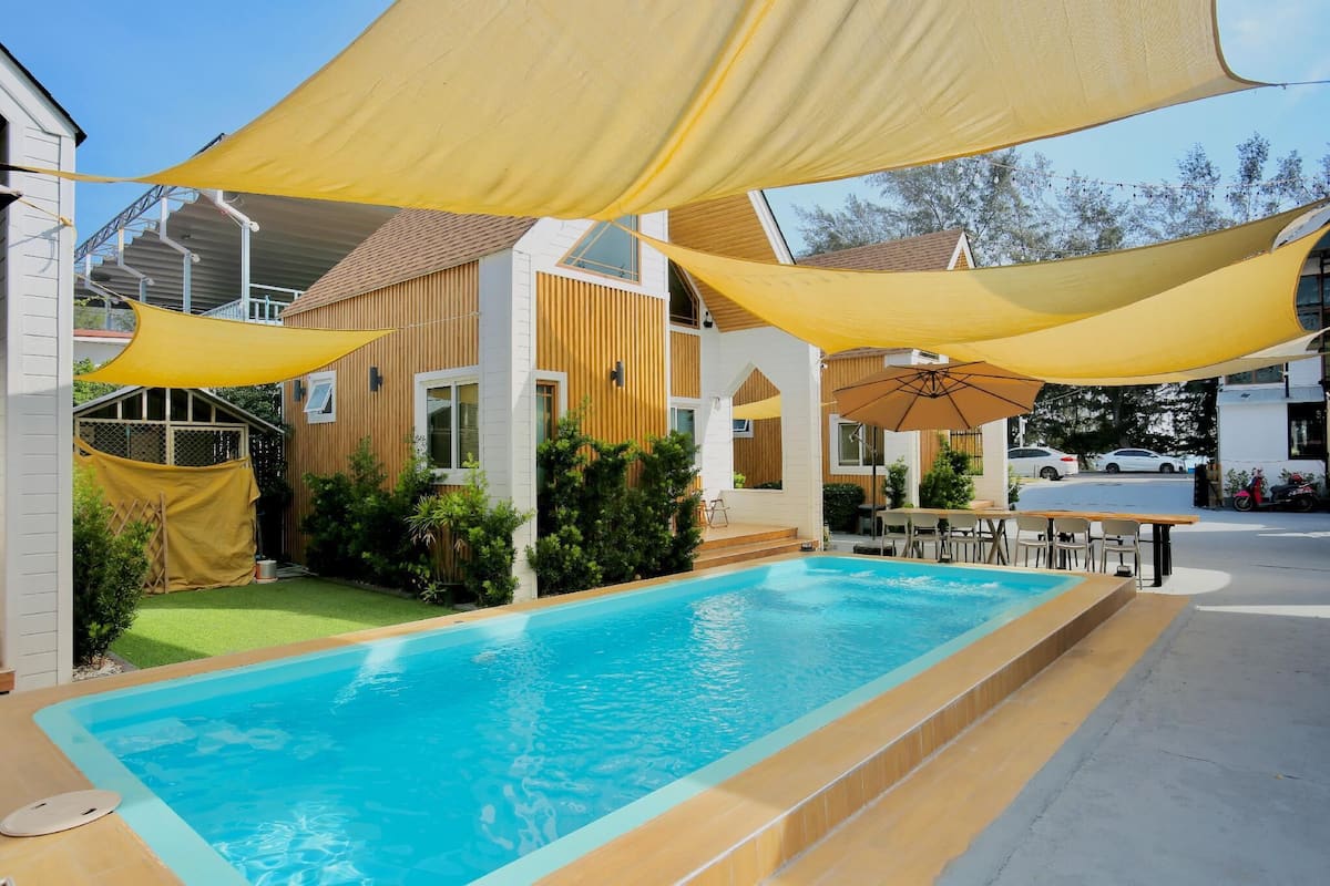 Piscina com toldo, área de estar coberta e um edifício com telhado inclinado.