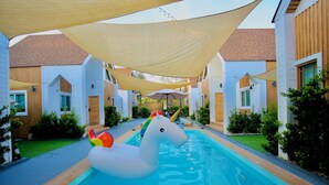 A pool area with a white inflatable unicorn float, surrounded by white buildings with wooden accents and covered by large shade sails. - Moon in 9 haus Cafe & Villas (Rayong)