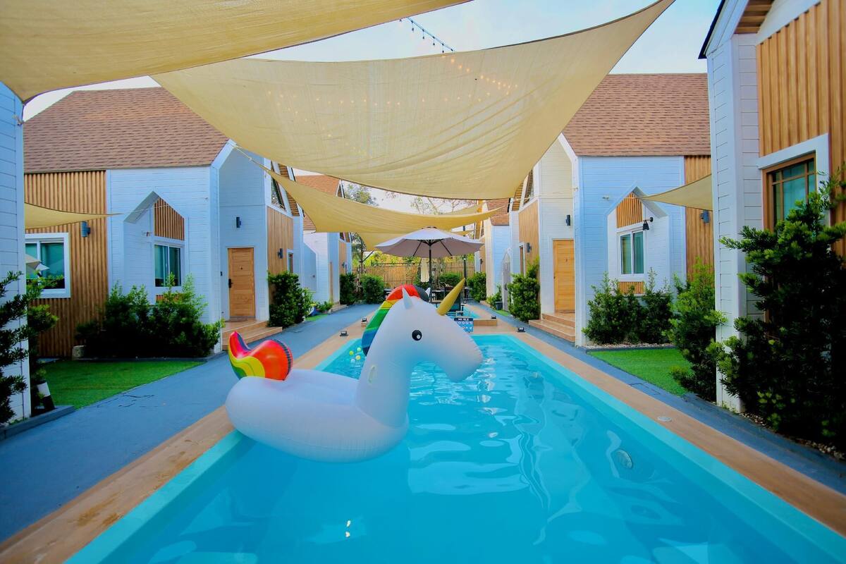 Uma área de piscina com um flutuador de unicórnio inflável branco, cercada por edifícios brancos com detalhes em madeira e coberta por grandes toldos de proteção.