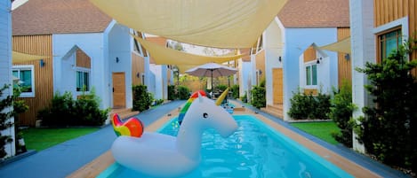Zona de piscina con un flotador inflable de unicornio blanco, rodeada de edificios blancos con detalles de madera y cubierta por grandes toldos de sombra.