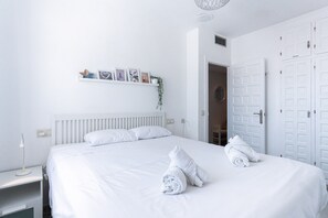 Habitación de hotel con una cama grande, ropa de cama blanca, una mesita de noche, un estante de pared con imágenes, y una puerta con manija.