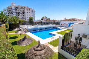 A swimming pool surrounded by a white building with balconies, a small garden with palm trees, and thatched umbrellas.