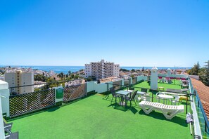 A rooftop terrace with green artificial turf, outdoor furniture, and a view of the sea and cityscape.