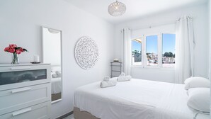 A white bedroom with a bed, a dresser, a mirror, and a window with a view of buildings. - Verano Azul Seasun 90B by Casasol (Nerja)