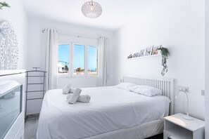 Een witte kamer met een bed, een raam, een radiator en een wandplank met foto's.