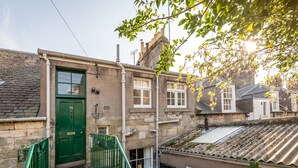 A narrow alleyway with a green door, stone walls, and a small staircase leading to a building with a tiled roof.