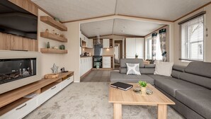 Ein modernes Wohnzimmer mit Kamin, einer Couch, einem Couchtisch und einem Fernseher. Im Hintergrund ist eine Küche zu sehen.