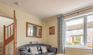 A living room with a leather sofa, a wooden staircase, a large window with a view of a residential area, and framed artwork on the wall.