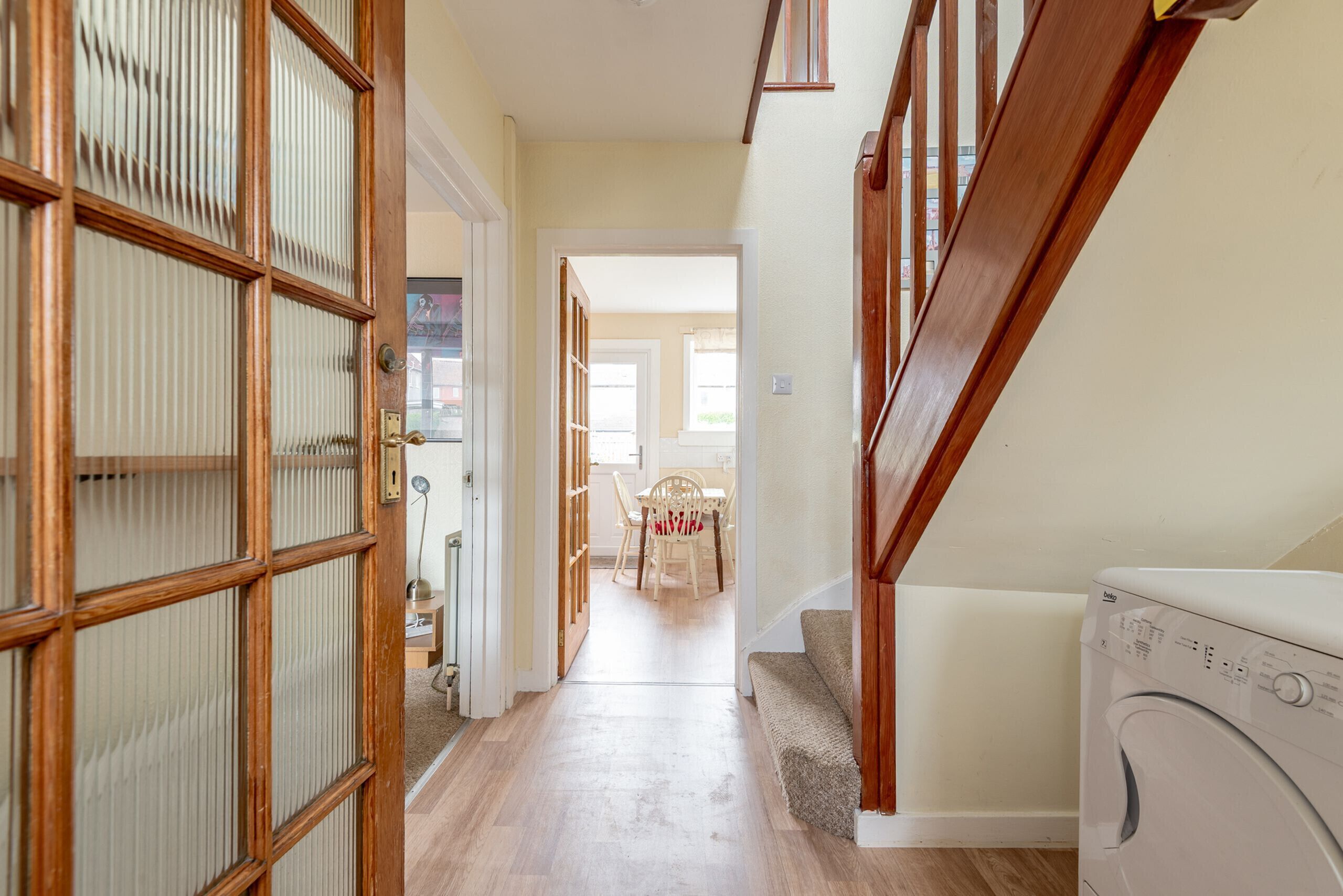 A hallway with a wooden staircase, a washing machine, and a dining area in the background.
