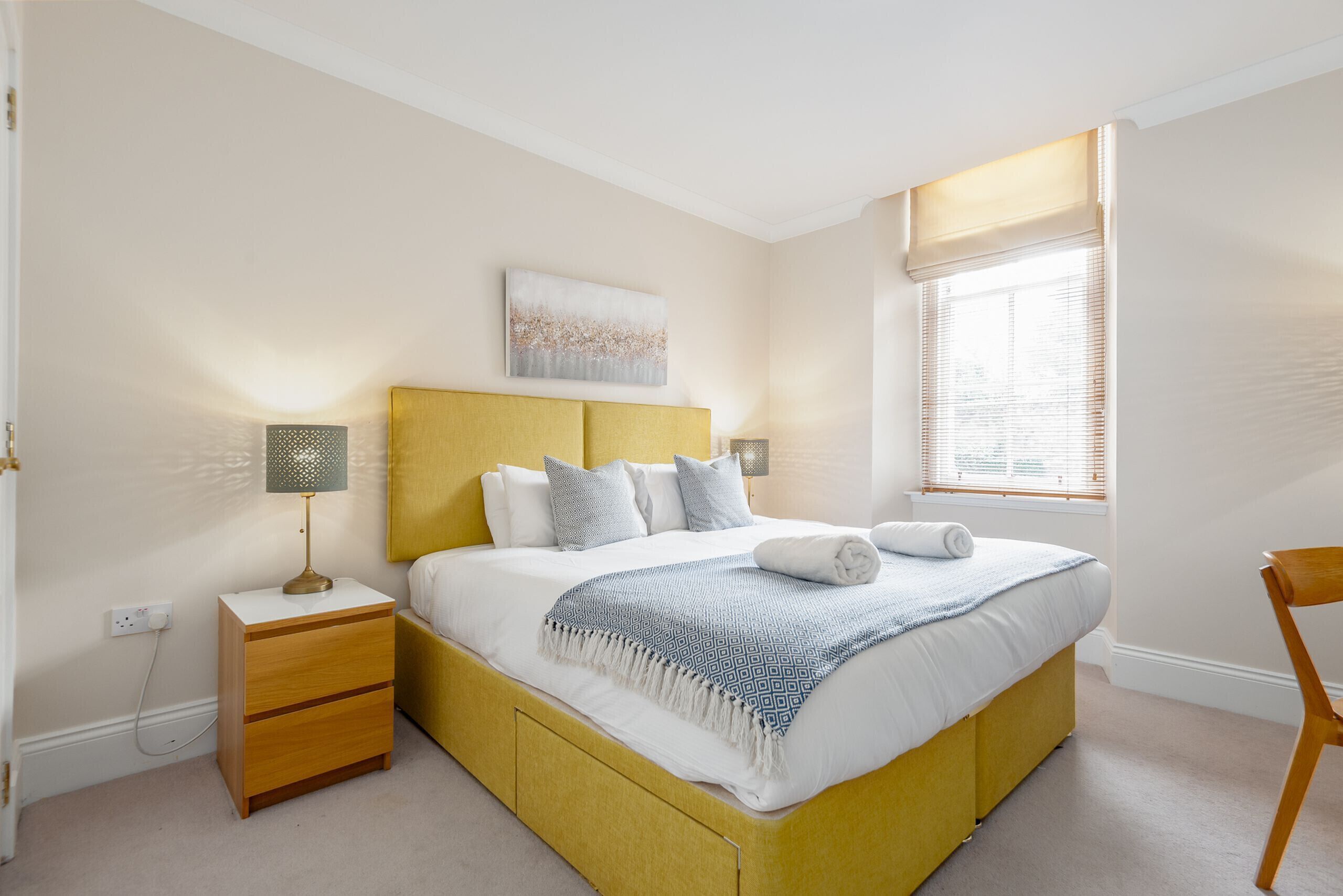 A bedroom with a yellow bed, white bedding, a bedside lamp, and a window with blinds.